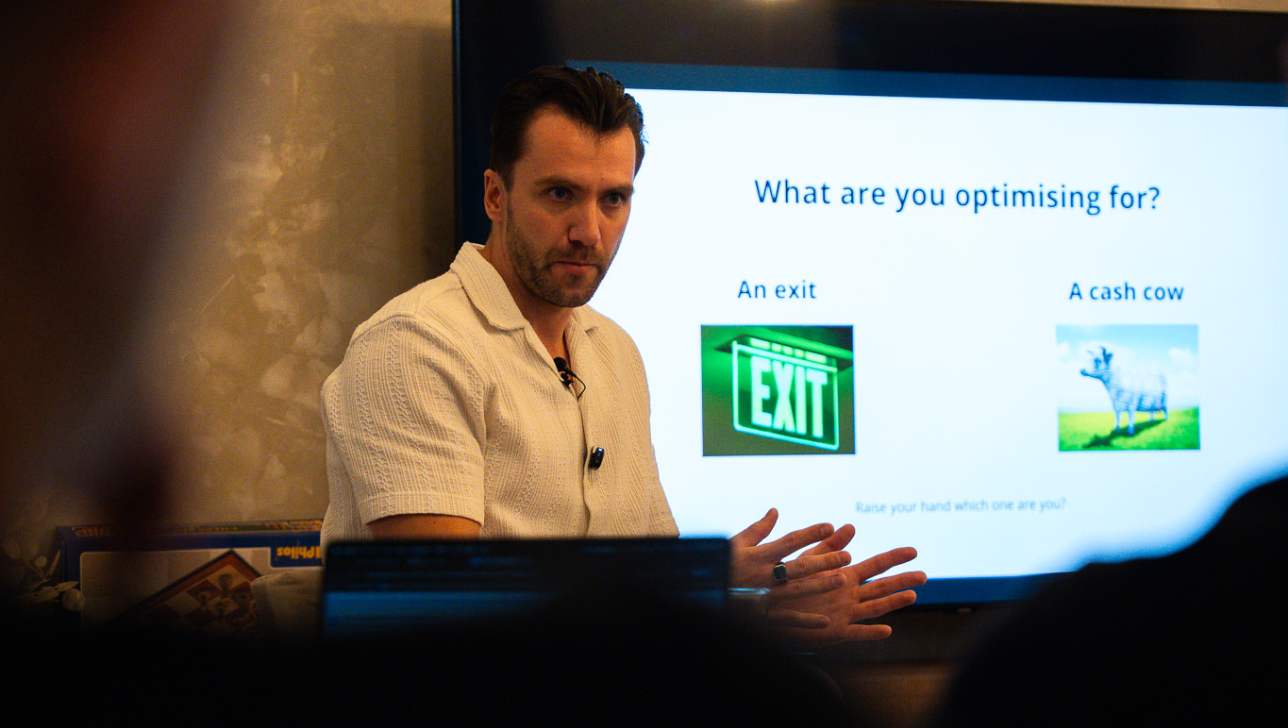 Romans Ivanovs at a BGG Agency Mastermind session, with the slide 'What are you optimising for?' on screen behind him.